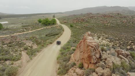 el avión no tripulado sube lentamente hacia arriba junto a una carretera donde un vehículo fuera de carretera negro conduce en el área silvestre de cederberg en sudáfrica - en el fondo se pueden ver montañas rocosas