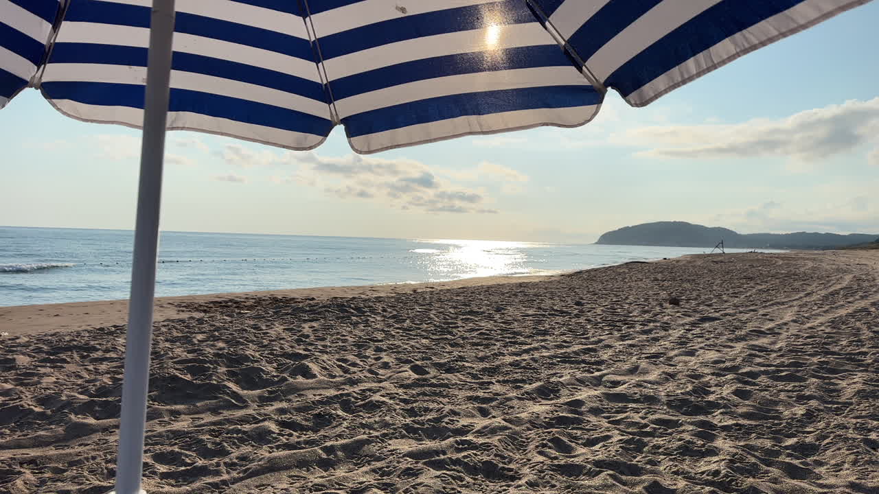 Scenic view from beneath a striped beach umbrella, overlooking a sandy shoreline and calm sea under the sunlight. A peaceful and relaxing coastal scene perfect for vacation, summer, and leisure themes