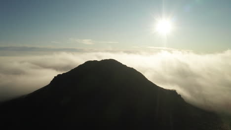 mountaintop peaking high above fog and clouds with bright sun, aerial sideways