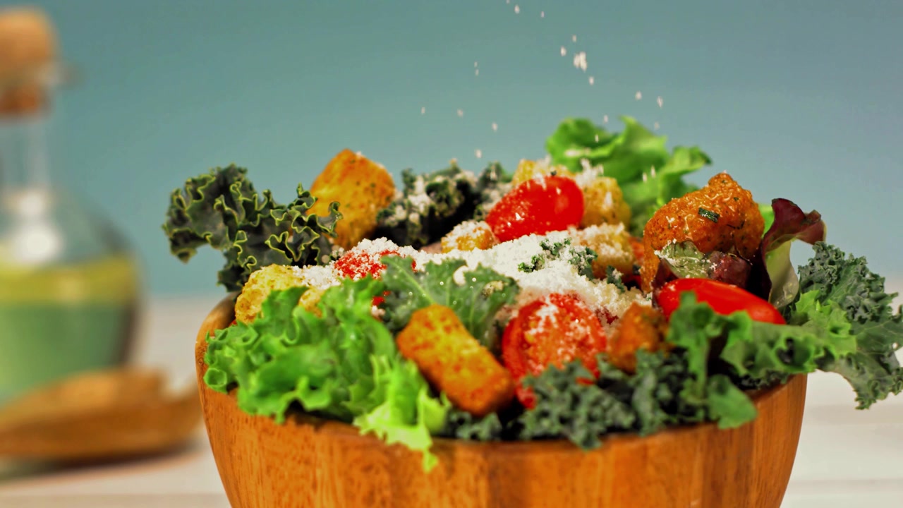 Slow motion close-up of grated Parmesan falling over a fresh green salad with lettuce, kale, cherry tomatoes and croutons in a rustic wooden bowl.