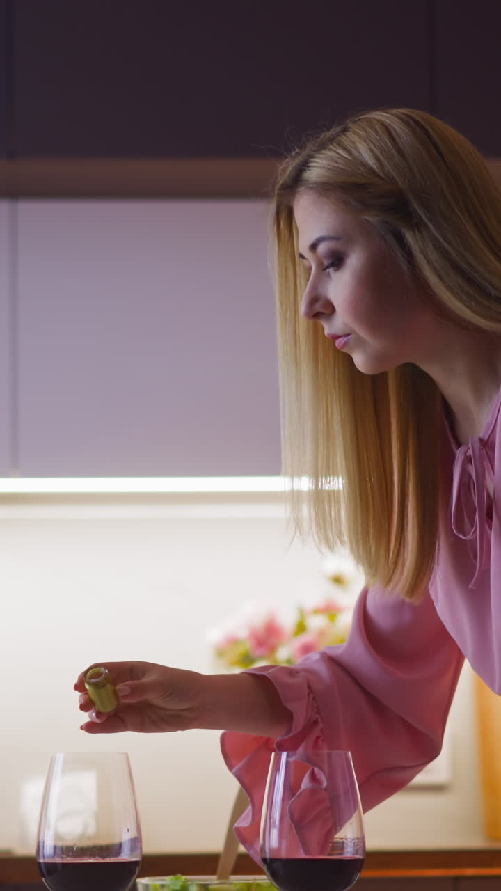 una mujer rubia pérfida agrega polvo venenoso al vino rojo de un visitante y mira a la cámara antes de cenar en una cocina elegante en cámara lenta.