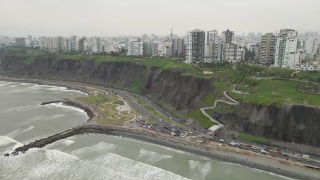 Esplendor-Panorámico-En-Miraflores:-Un-Dron-Captura-La-Sinergia-De-Las-Olas-Del-Océano,-La-Carretera-Costera-Y-El-Horizonte-Urbano