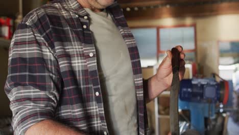 Midsection-of-caucasian-male-knife-maker-in-workshop,-holding-and-checking-knife