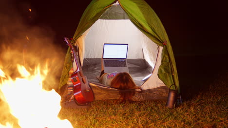 young girl chatting with laptop inside her tent while camping outdoor at night in front at bonfire