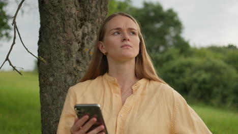 Young--Woman-is-thinking-and-holding-phone-outdoors