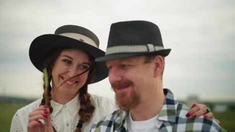 a close-up of a loving couple standing together, both wearing hats. the man, in a checkered shirt, is gently touched on the face by a piece of grass held by the woman in a white dress