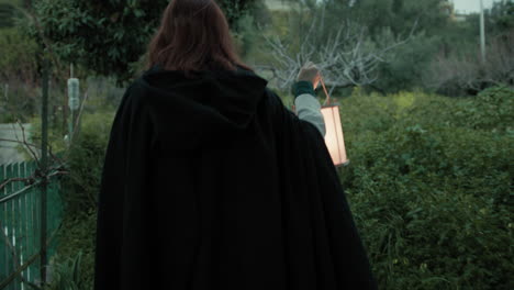 woman with cloak walks suspiciously among plants and trees with a lantern