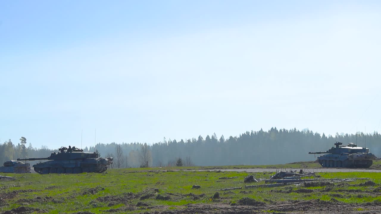 Multiple Challenger 2 4034 tanks looking for enemies on a grassy green and smoke covered summer field in front of a forest with their large cannon weapons at sunny day. Armed soldiers on top of tanks.