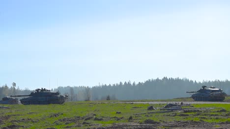 multiple challenger 2 4034 tanks looking for enemies on a grassy green and smoke covered summer field in front of a forest with their large cannon weapons at sunny day. armed soldiers on top of tanks.