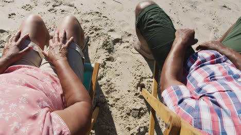 senior african american couple sitting on sunbeds and holding hands at the beach