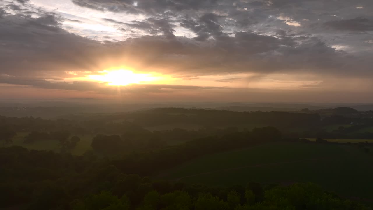 Sunrise shining over misty fields, woodland and hedgerows of South West England. Camera with slow altitude drop. Summer. Cornwall, UK.