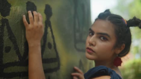 un primer plano en el rostro de una mujer joven de belleza india, de pie cerca de una pared de hormigón adornada con pinturas distintivas, inclinando la cabeza hacia arriba cuando la cámara se acerca a su cara