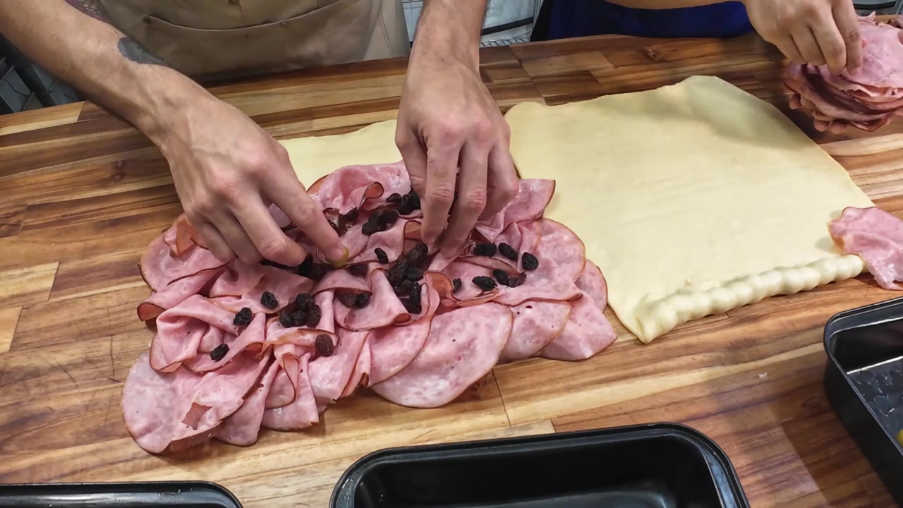Vista dall'alto di due panettieri che lavorano in sincronia, stendendo prosciutto e olive sull'impasto su un tavolo di legno, mostrando la preparazione professionale del cibo e il lavoro di squadra.