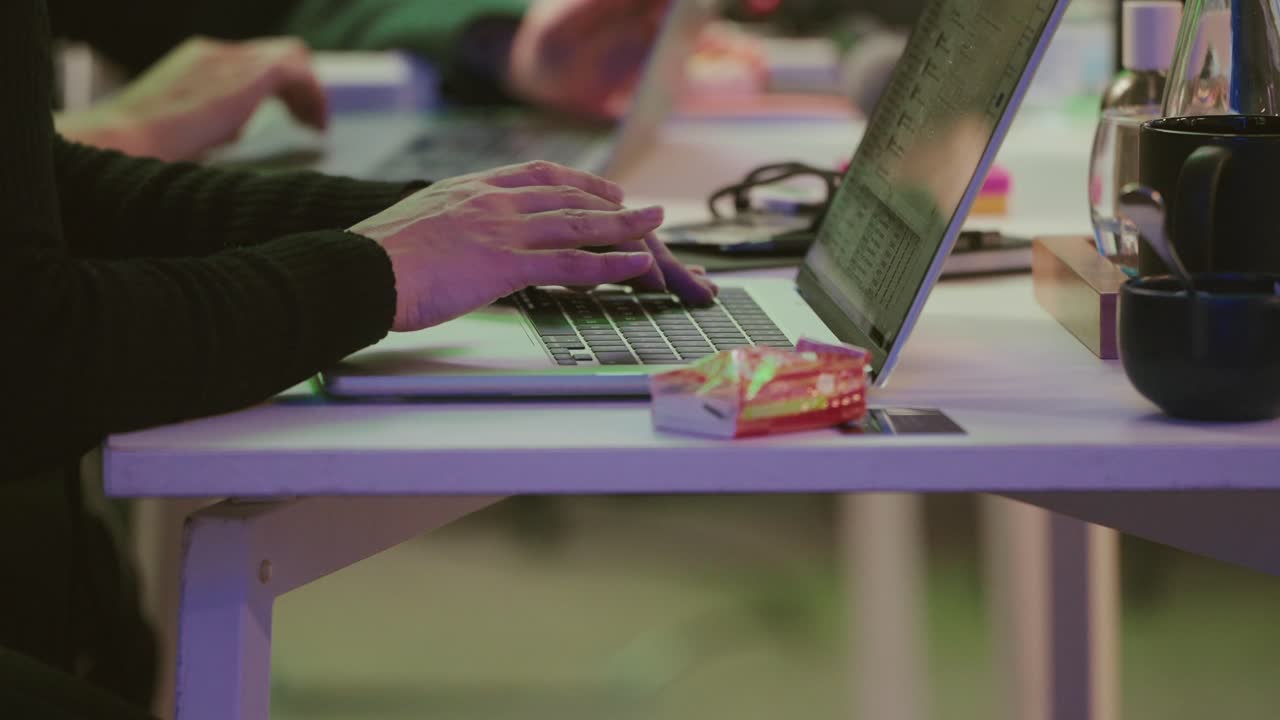 Person arbejder på bærbar computer ved et bord med kopper og slik i nærheden.