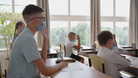 Kinder-In-Der-Schule-Sitzen-Im-Klassenzimmer-Und-Heben-Ihre-Hände,-Um-In-Zeitlupe-Die-Fragen-Des-Lehrers-In-Schutzmasken-Zu-Beantworten