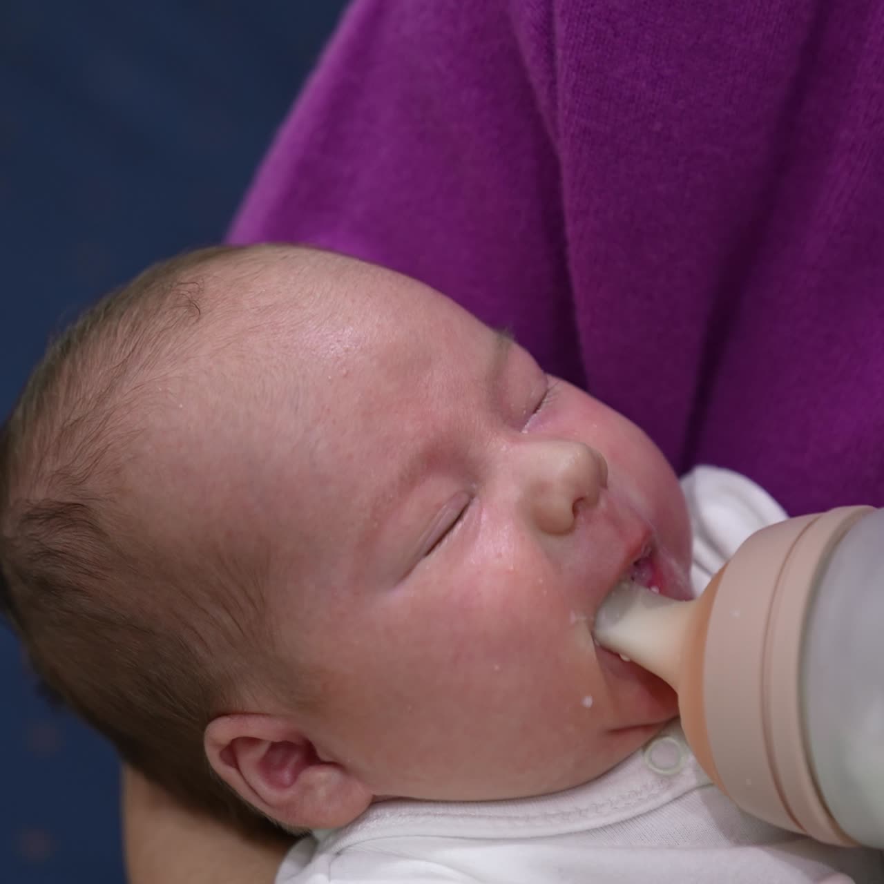 Little baby opens its mouth in hunger. Mother gives a bottle to her child, it suckles and milk drops appear on its cheek. Close up