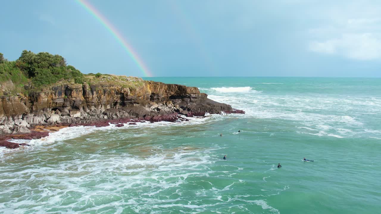toma de drones de las olas rompiendo los acantilados en la playa, con un arco iris en el fondo