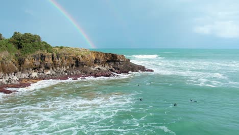 Toma-De-Drones-De-Las-Olas-Rompiendo-Los-Acantilados-En-La-Playa,-Con-Un-Arco-Iris-En-El-Fondo