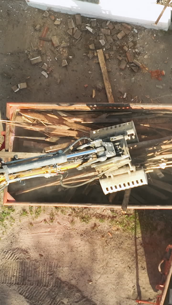 Top down aerial view of a hydraulic excavator claw sorting and loading scrap wood into a metal container. Concept for sustainable construction waste management and ecofriendly recycling