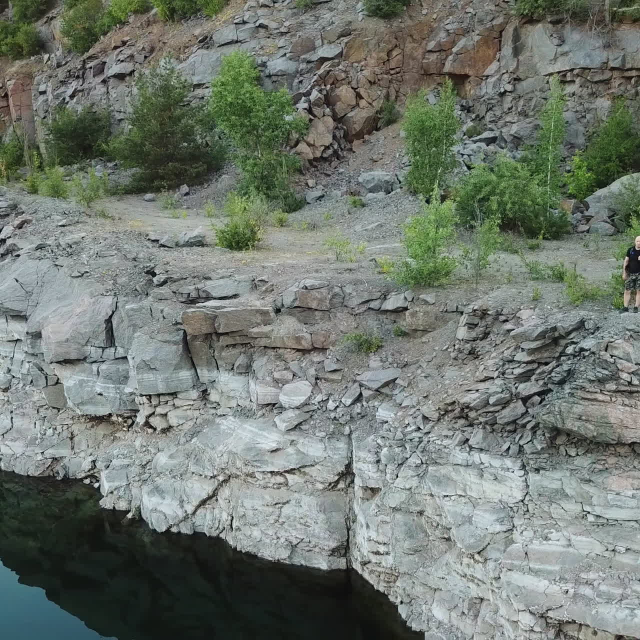 Young tourist man standing on steep slope with backpack over the water. Camper observing beautiful view of canyon with green trees and bushes and their reflection in the water. Aerial view