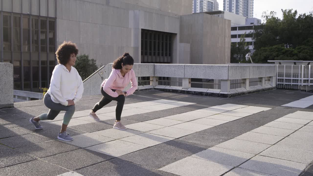 Two female friends starting workout with side stretches, pausing phone then jogging across rooftop