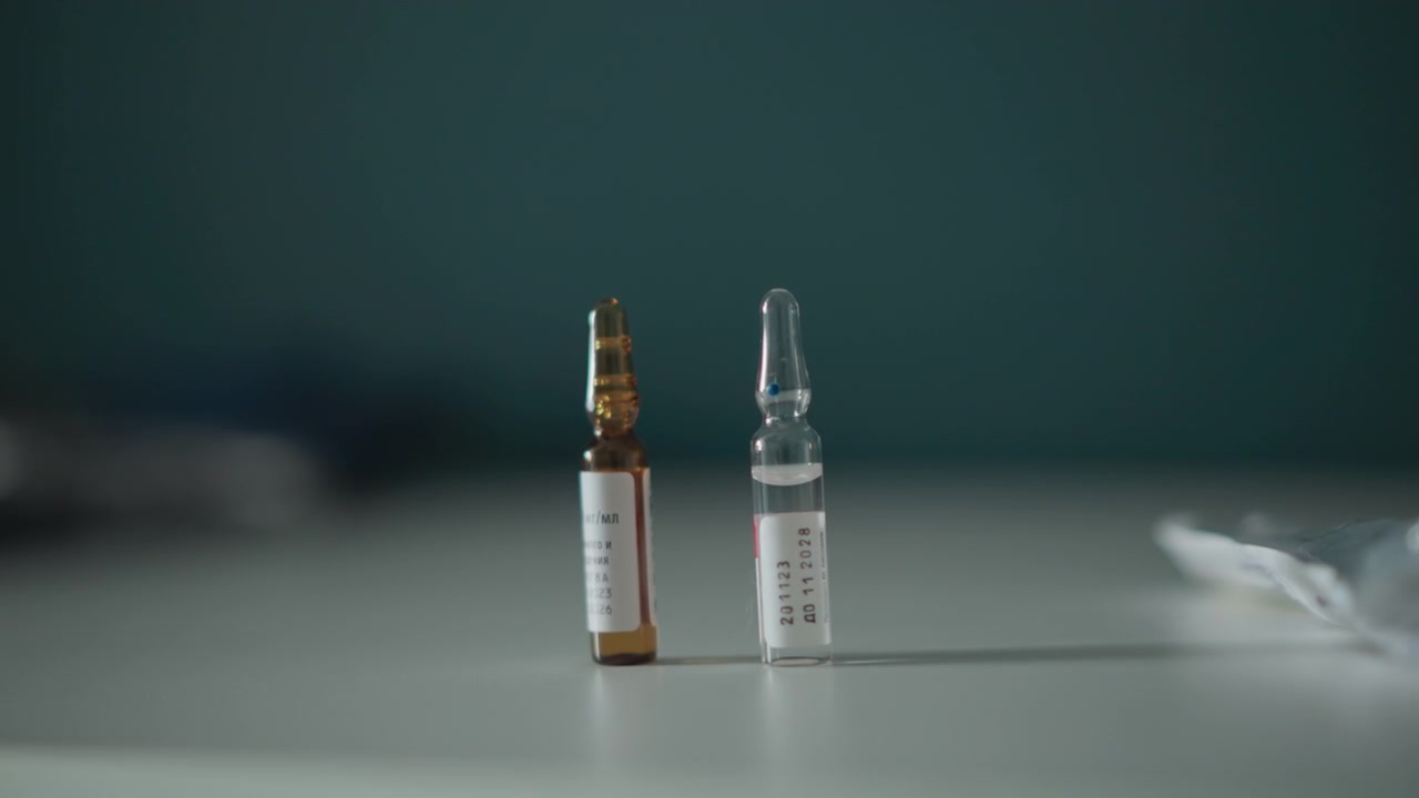 Mano guantata che posiziona una fiala ambrata sul tavolo, primo piano di fiale di vetro ed etichette, operatore sanitario che prepara il set di farmaci per l'iniezione, piano di lavoro sterile e azione di dosaggio precisa in un ambiente clinico calmo.