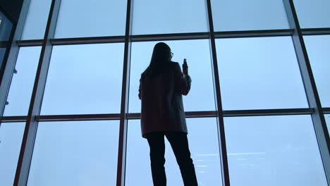 dark silhouette of a long-haired woman standing with her back. lady speaking on the phone in front of panoramic window. low angle view.