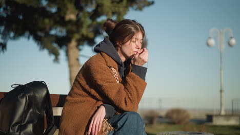 a girl sits alone on a bench in an urban park, overwhelmed with grief, her hand resting on her face. she wears a brown coat and blue jeans, with a black backpack placed beside her