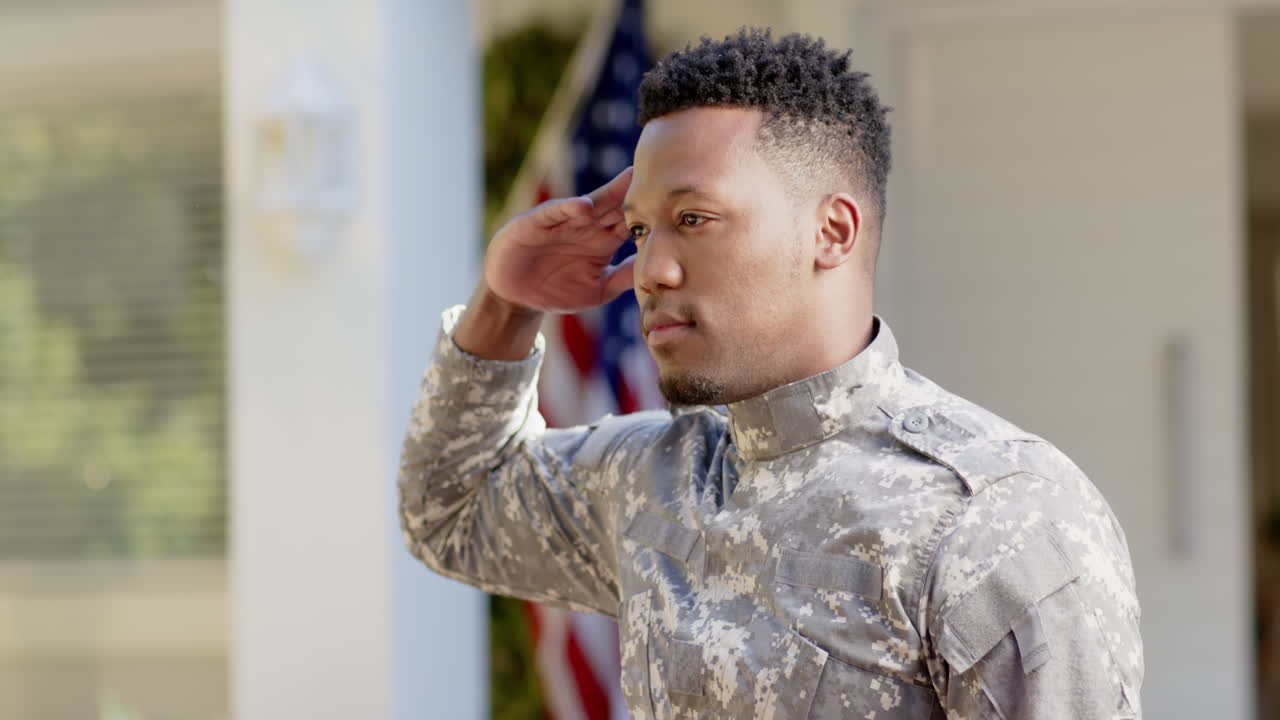 Happy african american male soldier saluting over flag of usa at home, slow motion
