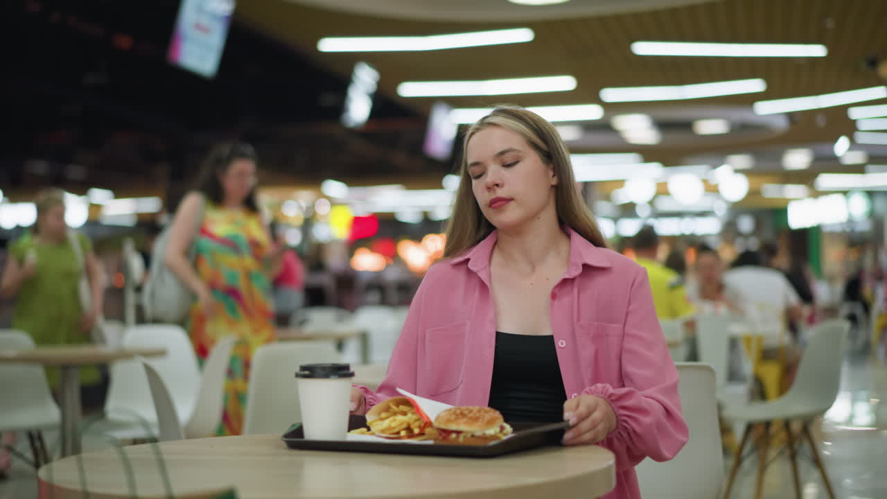 dama de vestido rosa se sienta en el restaurante, papas fritas, hamburguesa, y café en una bandeja, ella alcanza el café con iluminación bokeh y un fondo borroso lleno de gente moviéndose alrededor