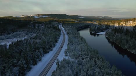 car driving alone on a country road surrounded by forest and along river, winter wonderland, aerial