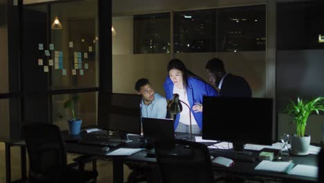 video of three happy diverse male and female colleagues using computers talking at night in office