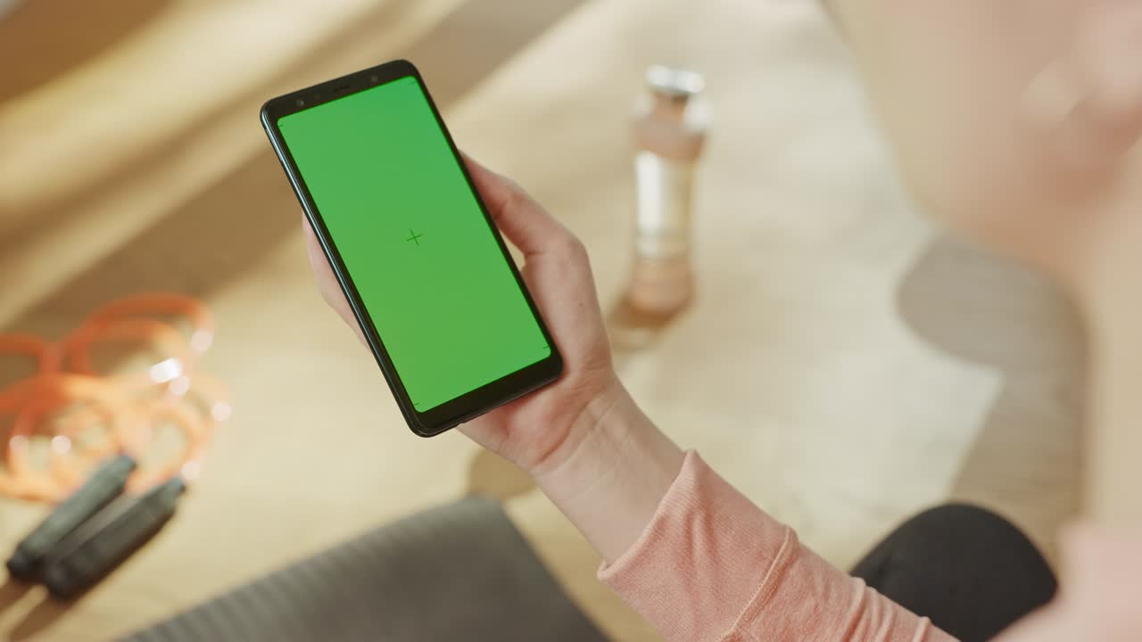 스 컨셉: 크로마 키를 들고 있는 여자, 초록색 스크린 스마트폰, 배경에는 피트니스 매트, 점프 로프, 장비, 근접 관점, 포브