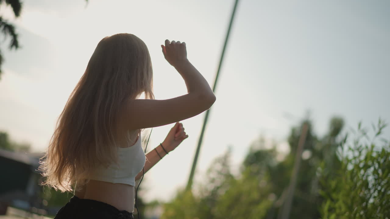 mulher branca girando em um pôr do sol suburbano, movimentos de freestyle energéticos com rodopio de cabelo e vento, silhueta retroiluminada por estrada e árvores, roupas casuais e fones de ouvido, brilho solar cinematográfico e quente