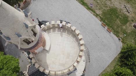 tourist man standing on a balcony at a european castle on top of a hill, green grass foliage - dolly drone shot