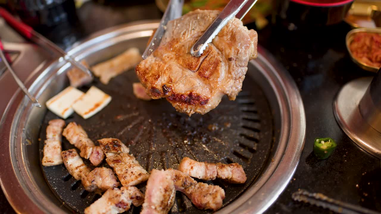 Hand uses tongs to flip pork on tabletop grill, surrounded by grilled meat and vegetables