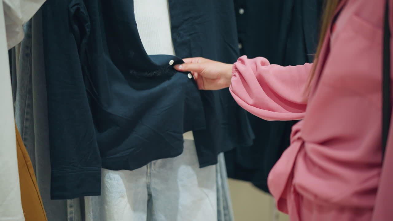 분홍색 셔츠를 입은 여자의 손은 랙에 있는 파란색과 색 옷을 만지기 위해 손을 고, 라벨을 확인하는 동안 직물의 질감을 느끼고, 랙에있는 다른 옷과 함께, 배경이 잘 밝습니다.