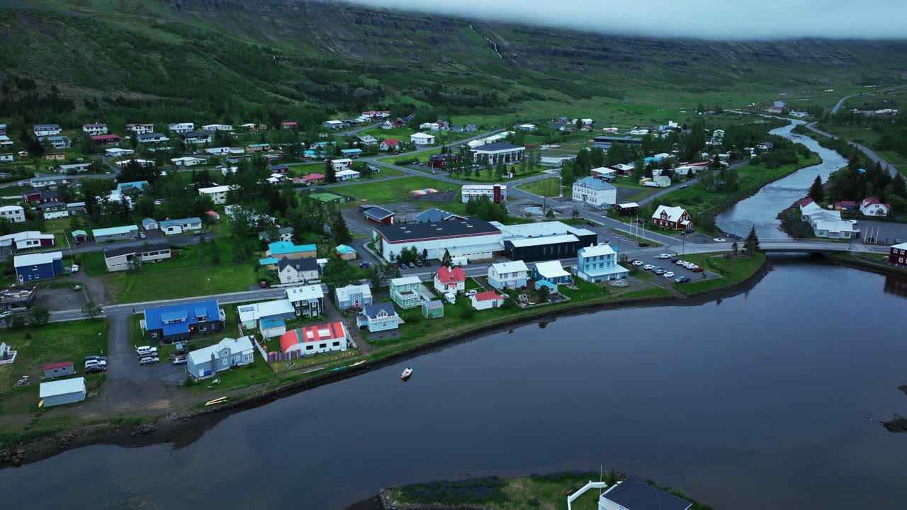 Icelandic settlement with colored houses and green mountains range. Aerial wide shot. Bridge and Fjord water in summer. Small neighborhood of Seyðisfjörður
