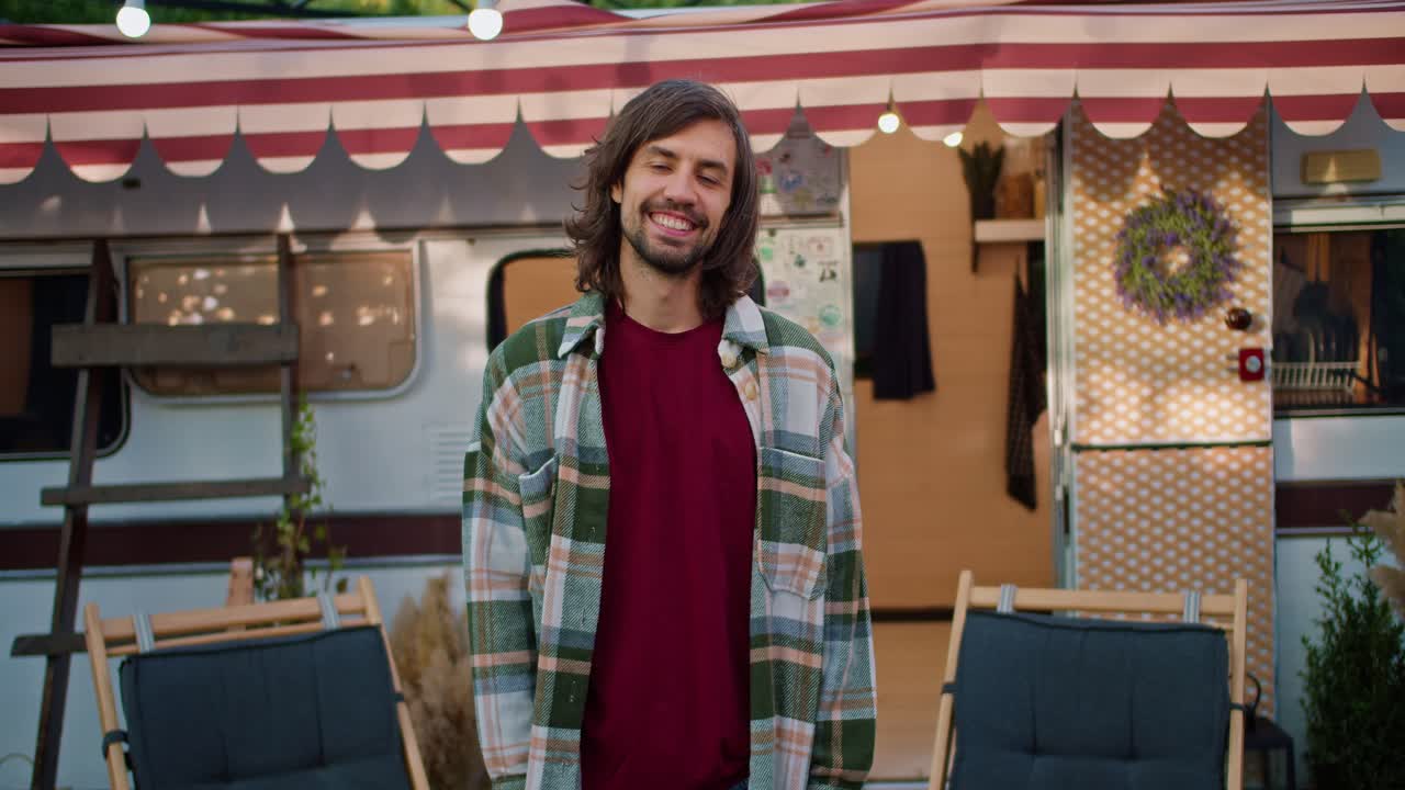 Portrait of a Happy brunette guy with stubble in a Green checkered shirt and a red T-shirt who stands near his decorated trailer during his picnic outside the city at a camp in the summer