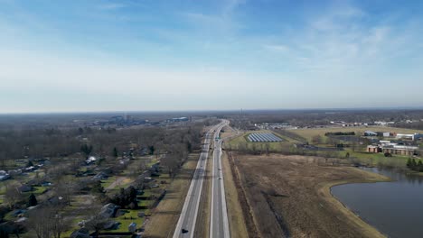 drone flying overhead a 2 lane highway with a small lake, solar panels, and college on the right hand side