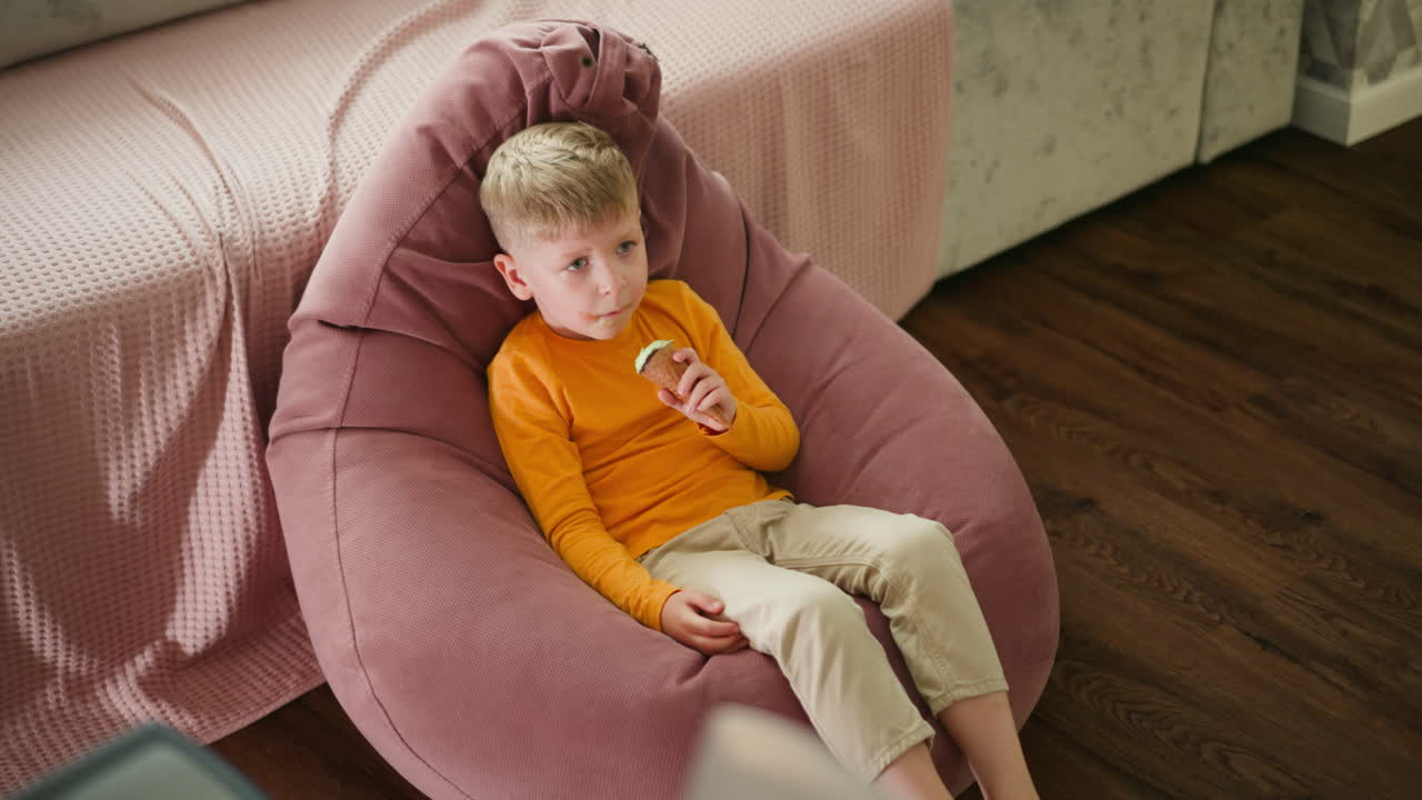 Aerial view of young white boy seated comfortably on pink beanbag indoors, enjoying ice cream cone while playfully moving legs, with wooden floor and soft furnishings around