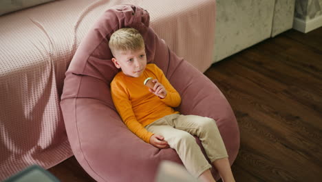 aerial view of young white boy seated comfortably on pink beanbag indoors, enjoying ice cream cone while playfully moving legs, with wooden floor and soft furnishings around
