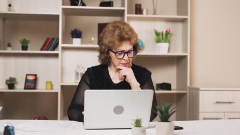 Die-Alte-Frau-Führt-Die-Bücher,-Sitzt-An-Ihrem-Laptop-Und-Macht-Notizen-Auf-Einem-Blatt-Papier