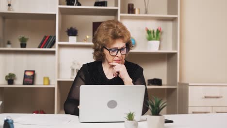 old woman is keeping the books, sitting at her laptop and jotting down on a piece of paper