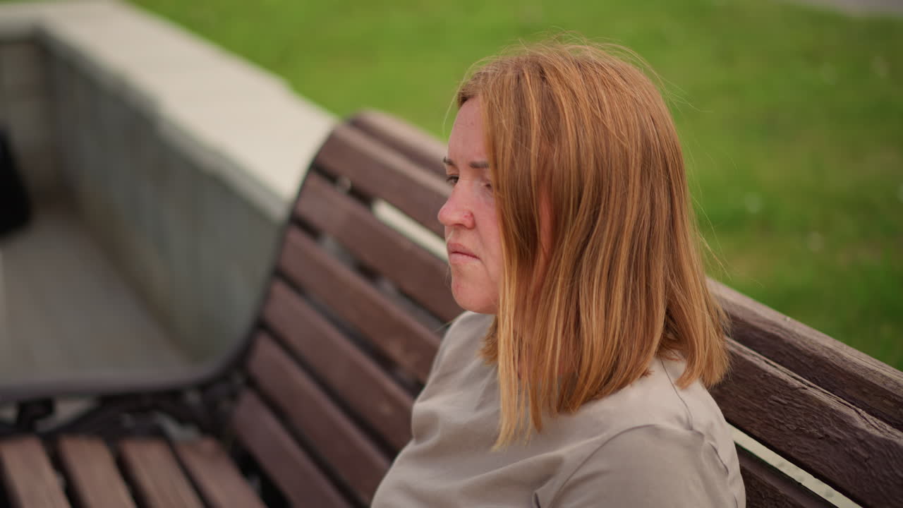 Crying woman sitting outdoors on wooden bench wiping tears with tissue as car passes in blurred background, emotional face expressing sadness and loneliness