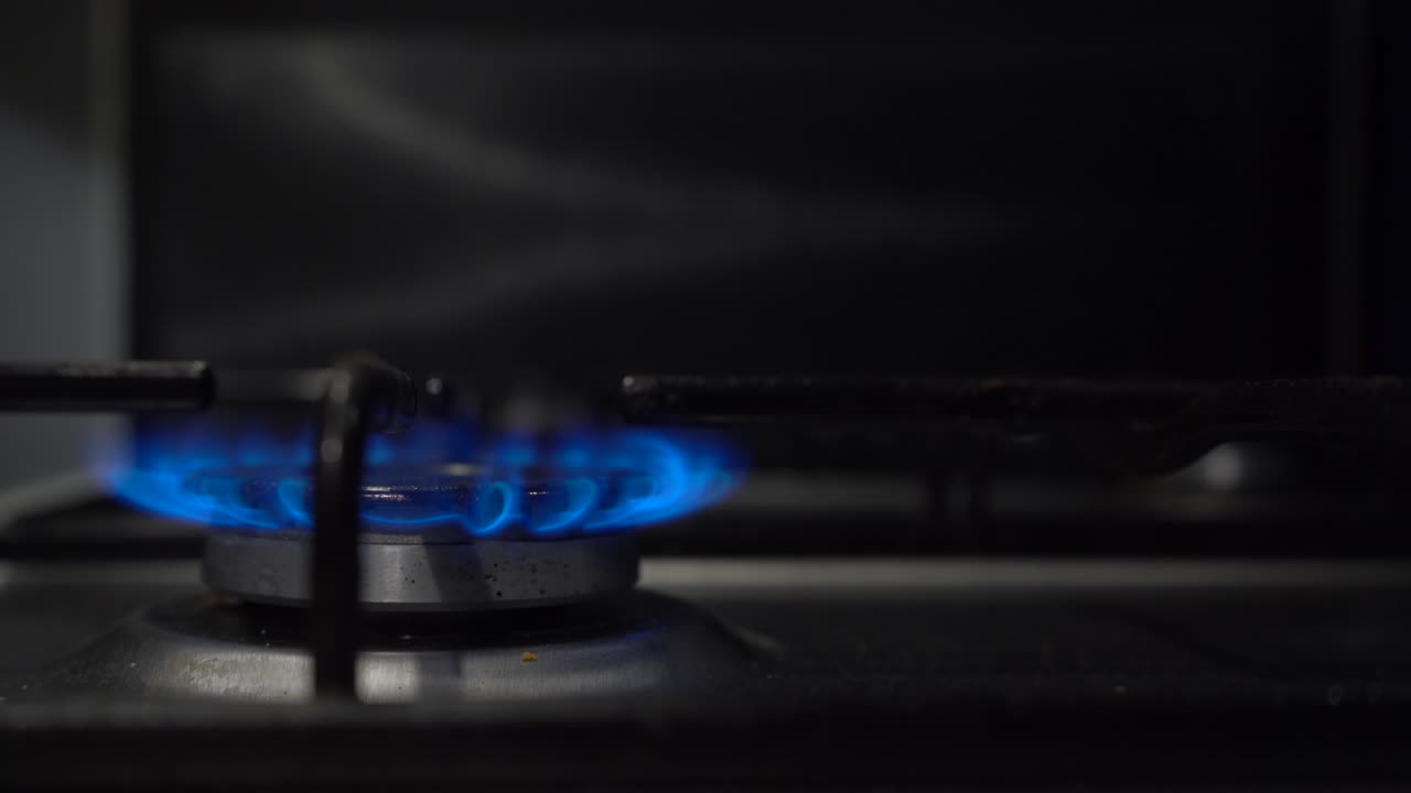 газовая плита горит синим пламенем, расфокусированная рука на заднем плане поворачивает ручку