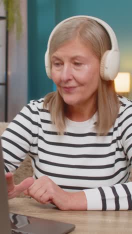 mature old woman listening to music on laptop with wireless headphones relaxed satisfied at home