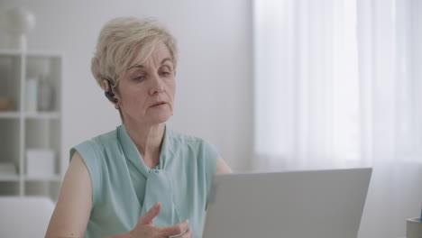 La-Especialista-Femenina-Del-Centro-De-Llamadas-Está-Hablando-Con-Auriculares-Y-Mirando-La-Pantalla-Del-Retrato-De-Una-Computadora-Portátil-En-El-Interior-Trabajando-De-Forma-Remota