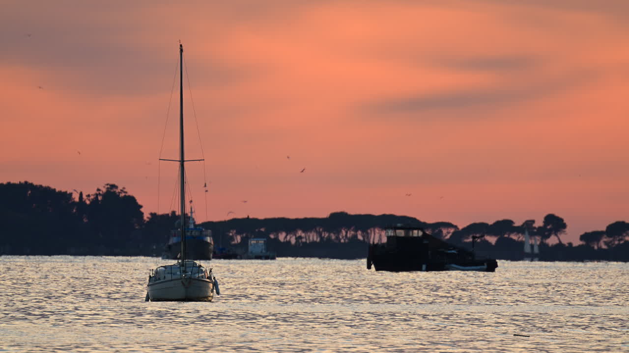 Piccole imbarcazioni ancorate al largo durante il tramonto, con calde tonalità arancioni e rosa che si riflettono sul mare e silhouette di alberi in lontananza
