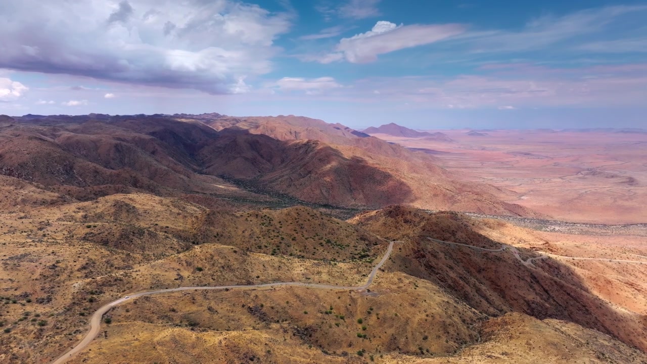 Veduta aerea di montagne aride e piegate che scendono verso le ampie pianure desertiche del Namib. Una solitaria strada sterrata si snoda attraverso le drammatiche montagne arse dal sole della grande scarpata.